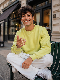 Realistic treet portrait, handsome young man with fluffy dark curly hair, sitting cross-legged on a dark green outdoor metal bench on city sidewalk, big bright smile showing teeth, right hand raised gesturing as if chatting, wearing lime yellow crew neck sweatshirt, loose white casual trousers, white sneakers, modern urban building background with shallow depth of field, natural soft daylight, lifelike skin texture, casual lifestyle photography, shot on Canon R5, 50mm f/1.8 lens, sharp focus, 8K, ultra detailed, warm natural tone