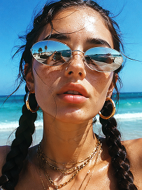 Close-up beach portrait, young woman with two thick dark long braids, freckles all over nose and cheeks, futuristic silver oval mirrored sunglasses reflecting beach scenery, large gold hoop earrings, layered multiple gold chain necklaces, slightly open mouth, bright harsh summer midday sunlight, clear turquoise ocean and blue sky background, wind blowing stray hair strands on face, realistic skin texture, sun-kissed complexion, vintage film grain, summer coastal fashion photography, sharp details, high saturation, shot on 35mm film, 8K ultra realistic